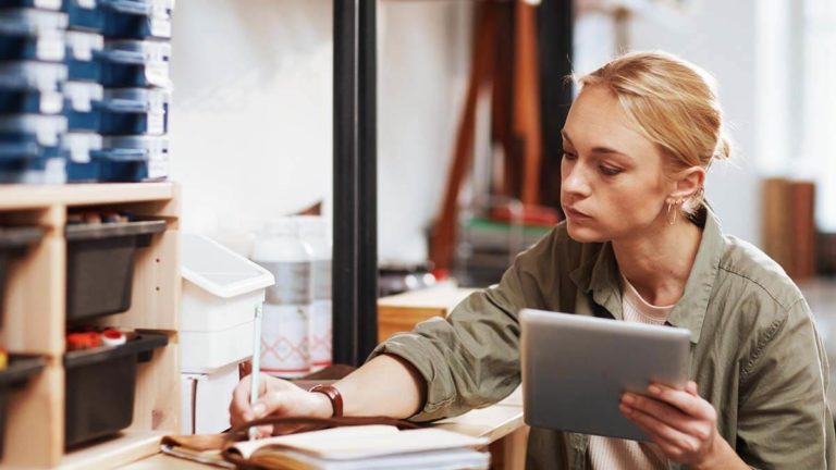 Woman holding a tablet and writing in a notebook.