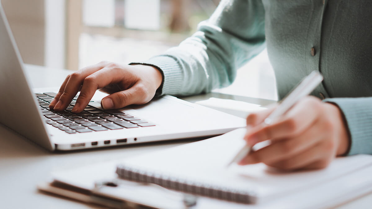 Business owner writing on spiral notepad and using laptop.