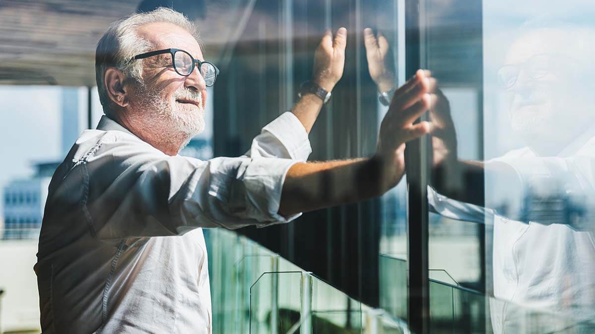 Older man leaning against glass windows and looking out at city.