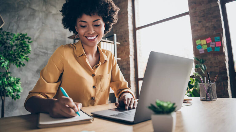 Smiling woman writing in a notebook and using a laptop.