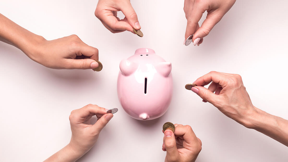 Six hands in a circle putting coins in a piggy bank.